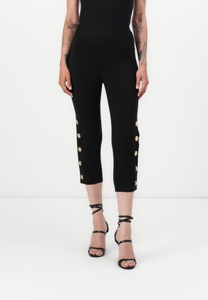 Black cropped pants with gold button accents on the sides, made from stretchy fabric. Paired with black strappy sandals.