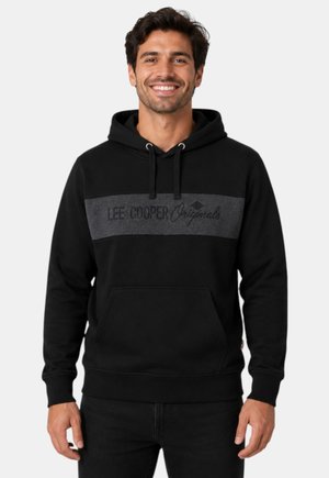 Hombre sonriente con una sudadera negra con capucha con un panel gris oscuro y el texto "Lee Cooper Originals" en el pecho, de pie frente a un fondo blanco.