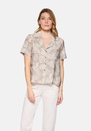 Mujer con una camisa beige de manga corta con botones y motivos blancos de hojas, y pantalones blancos, de pie frente a un fondo claro y sencillo.