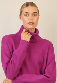 Magenta Rollkragenpullover mit lässiger Passform, gerippten Bündchen und weicher Textur. Das Modell greift leicht den Kragen und zeigt Details des Ausschnitts.