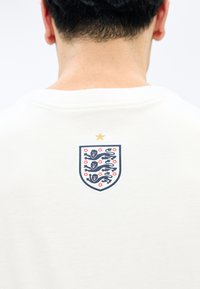 Άνδρας με κοντά μαύρα μαλλιά, φοράει λευκό πουκάμισο με το έμβλημα της ποδοσφαιρικής ομάδας της Αγγλίας, με τρεις λέοντες και ένα χρυσό αστέρι στην πλάτη.