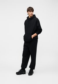 Μαύρο φούτερ με κουκούλα και μπροστινή τσέπη, σε συνδυασμό με μαύρο παντελόνι άνετης εφαρμογής και μαύρα sneakers με υφή. Απλός, μονόχρωμος σχεδιασμός.