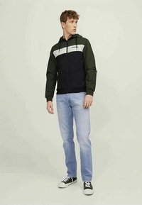Casaco desportivo com fecho éclair, capuz e mangas verdes, corpo preto e uma faixa cinza ao longo do peito. Combinado com jeans azul claro e ténis pretos.
