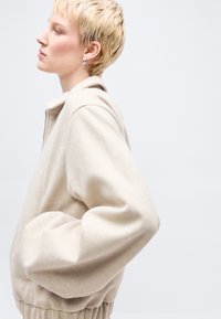 Persoană cu păr blond scurt, purtând cercei argintii și o jachetă bej, stând în profil pe un fundal deschis simplu.