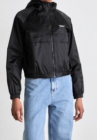 Svart lättviktsjacka med huva, elastiska muddar och midja, med en glansig finish och en logotyp på bröstet. Bärs tillsammans med ljusblå jeans.