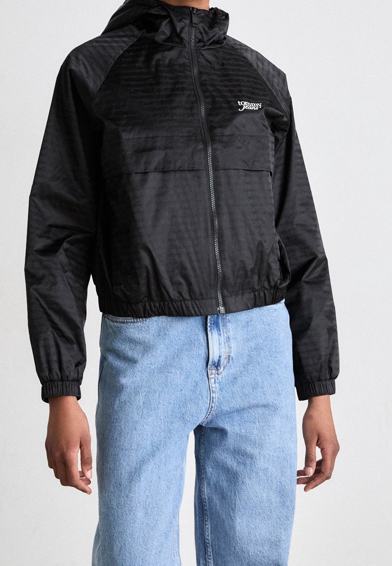 Svart lättviktsjacka med huva, elastiska muddar och midja, med en glansig finish och en logotyp på bröstet. Bärs tillsammans med ljusblå jeans.