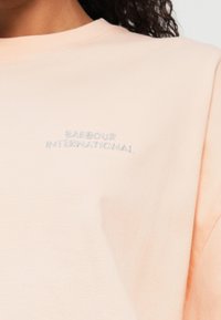 T-shirt de manga curta pêssego claro, feito de algodão macio, apresentando um discreto logo bordado "Barbour International" no peito.