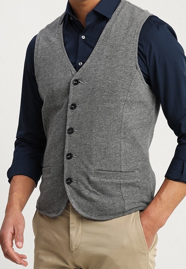 Homme portant un gilet gris boutonné sur une chemise bleu foncé aux manches retroussées et un pantalon beige, une main dans la poche.