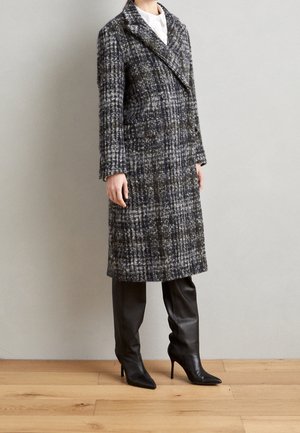 Femme portant un manteau gris à carreaux mi-long croisé et des bottes en cuir noires à talons montant jusqu'aux genoux, debout sur un sol en bois.
