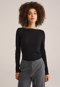Vrouw met krullend haar, gekleed in gouden oorringen, een zwart langemouwen top en grijze broek met trekkoord, staand tegen een beige achtergrond.