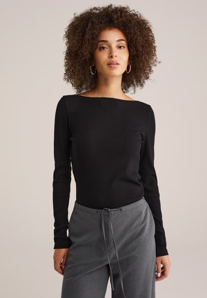 Vrouw met krullend haar, gekleed in gouden oorringen, een zwart langemouwen top en grijze broek met trekkoord, staand tegen een beige achtergrond.