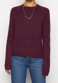 Pull en laine bordeaux à manches longues, col rond et texture douce, associé à un jean en denim bleu. Présenté sur un fond de couleur neutre.