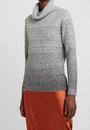 Personne portant un pull à col roulé gris à motifs et une jupe orange, vue des épaules jusqu'à mi-cuisse sur un fond uni.