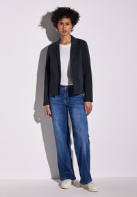 Svart blazer med en enda knapp, bärs över en vit t-shirt och blå utsvängda jeans. Vita sneakers fulländar outfiten. Slät, skräddarsydd tyg.