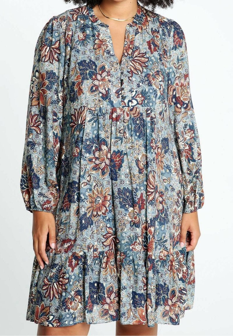 Robe à manches longues à motif floral bleu, rouille et beige, avec un décolleté en V et une taille froncée portée par une personne à la peau moyennement foncée.