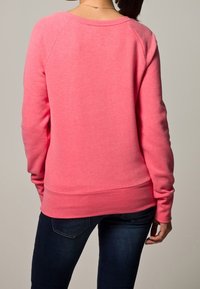 Heldroze sweatshirt met een ronde halslijn en ribbels aan de mouwen. Zachte stof met een gladde textuur. Slim fit, gedragen met donkerblauwe jeans.