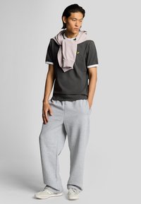 T-shirt cinza com detalhes brancos, sobreposta por um hoodie rosa claro aos ombros, combinado com calças de moletom cinzas largas e sapatos brancos.