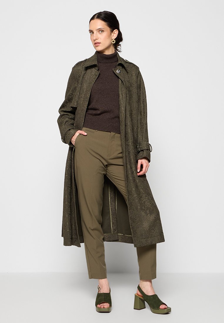 Cappotto lungo verde oliva con texture floreale, dolcevita marrone e pantaloni abbinati verde oliva. Indossa sandali con tacco verde con cinturino alla caviglia.
