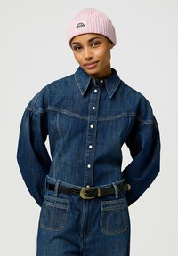 Camicia di denim con chiusura a bottoni e maniche oversize, abbinata a una cintura nera con una fibbia dorata. Un morbido berretto rosa a coste completa il look.