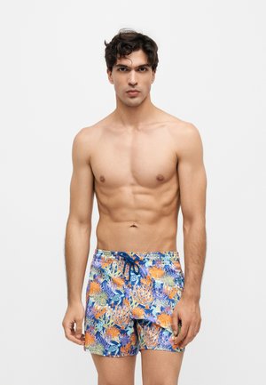 Jeune homme aux cheveux foncés portant un short de bain coloré à motif floral sur un fond clair uni.