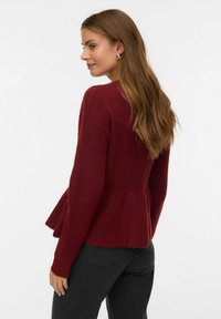 Burgundijska peplum pulover iz teksturirane pletene tkanine, z dolgimi rokavi in razširjenim spodnjim robom. Obučen z temnimi kavbojkami.