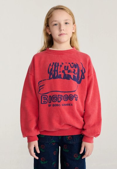 Enfant aux longs cheveux blonds porte un sweat-shirt rouge avec un imprimé bleu "BIGFOOT by BOBO CHOSES" et un pantalon foncé à motifs floraux rouges.