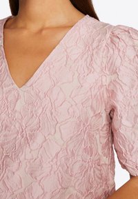 Różowa bluzka z haftem w kwiaty, z głębokim dekoltem w serek, teksturowanym materiałem i bufkowanymi rękawami, prezentująca skomplikowane wzory płatków na gładkim tle.