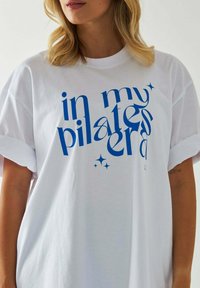 Biała bawełniana koszulka z oversize'owym krojem i podwiniętymi rękawami. Z przodu znajduje się niebieski napis "w mojej erze pilatesu" z akcentami gwiazdek.