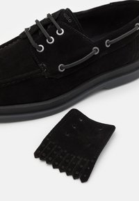 Schwarzer Wildlederschuh mit runder Zehenform, kontrastierenden grauen Ösen und einer flachen Gummisohle. Begleitet von einem passenden Akzentstück aus Wildleder.