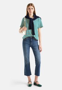 Mujer vestida con una camiseta a rayas verdes y blancas, jeans cortos azules, zapatos verdes, suéter azul marino sobre los hombros, sosteniendo un bolso blanco.