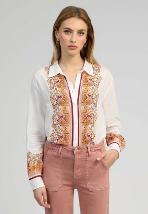 Jeune femme portant une blouse blanche à boutons avec une broderie florale orange et un pantalon rose taille haute, debout avec une main dans la poche.