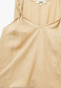 Beige ärmlös topp med V-ringning, med vridet detaljer vid axlarna, en ficka på vänster sida och en strukturerad tyg.