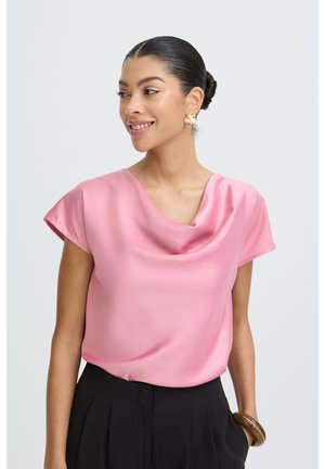 Femeie cu păr închis la culoare prins în coc, purtând o bluză roz din satin, pantaloni negri, cercei mari aurii și o brățară groasă de aur, zâmbind și privindu-se pe partea laterală.