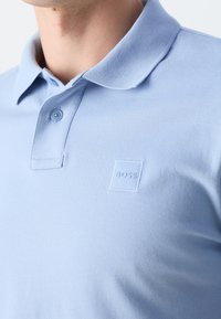Gros plan d'une personne portant un polo bleu clair avec un col, une patte de boutonnage et un petit logo en relief "BOSS" sur la poitrine.