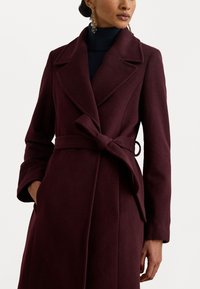 Cappotto in misto lana borgogna con vita svasata, ampie rever e maniche lunghe. Tessuto liscio con tasche laterali e vestibilità su misura.