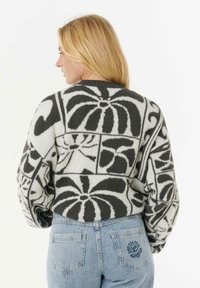 Donna bionda che indossa un maglione con motivo nero e bianco con disegni floreali astratti e jeans azzurri con ricami di sole e fiore.