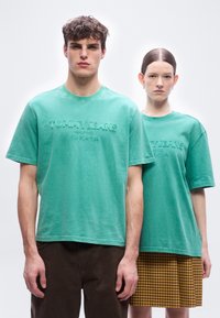 Άνδρας και γυναίκα στέκονται δίπλα-δίπλα φορώντας ταιριαστά τιρκουάζ t-shirts "Tommy Jeans". Ο άνδρας φοράει σκούρο παντελόνι, η γυναίκα φούστα κίτρινη καρό.