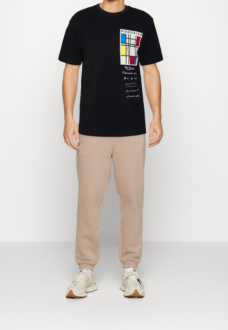 Sort bomulds-T-shirt med Mondrian-inspireret grafik i rød, gul og blå. Kombineret med beige joggingbukser og lyse sneakers.