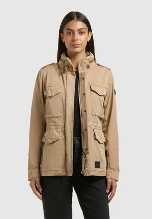 Nő bézs színű, négy zsebes, felhajtókupakkal és magas gallérral rendelkező munkakabátban, fekete nadrágot viselve, egyszínű háttér előtt áll.