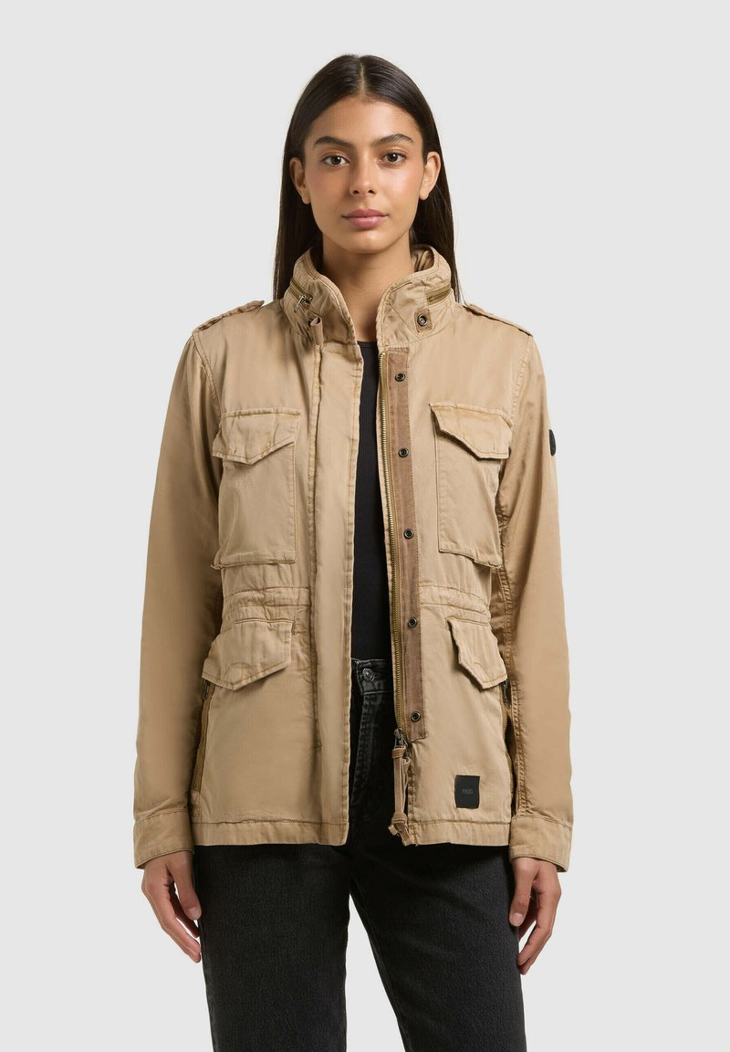 Donna che indossa una giacca utility beige con quattro tasche con patta e un colletto alto, abbinata a pantaloni neri, in piedi contro uno sfondo semplice.