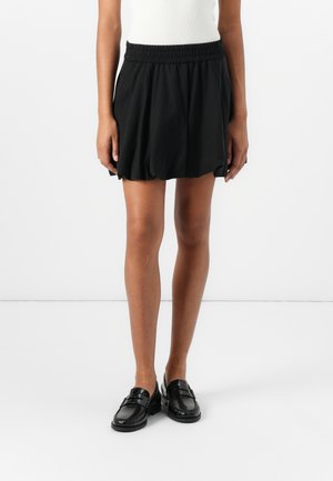 Person iført en sort elastisk talje bubble nederdel, kombineret med sorte læder loafers og en hvid top, stående mod en ensfarvet baggrund.