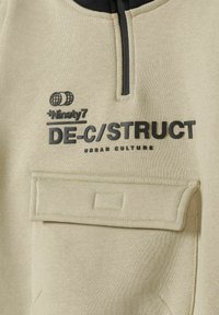 Light beige sweatshirt with black accents, half-zip neck, textured fabric, front pocket, and printed logos "Ninety7" and "DE-C/STRUCT."