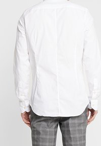 Biała koszula z długim rękawem o prostym kroju, z guzikami przy mankietach oraz środkowym szwem na plecach. Materiał wydaje się lekki i gładki.