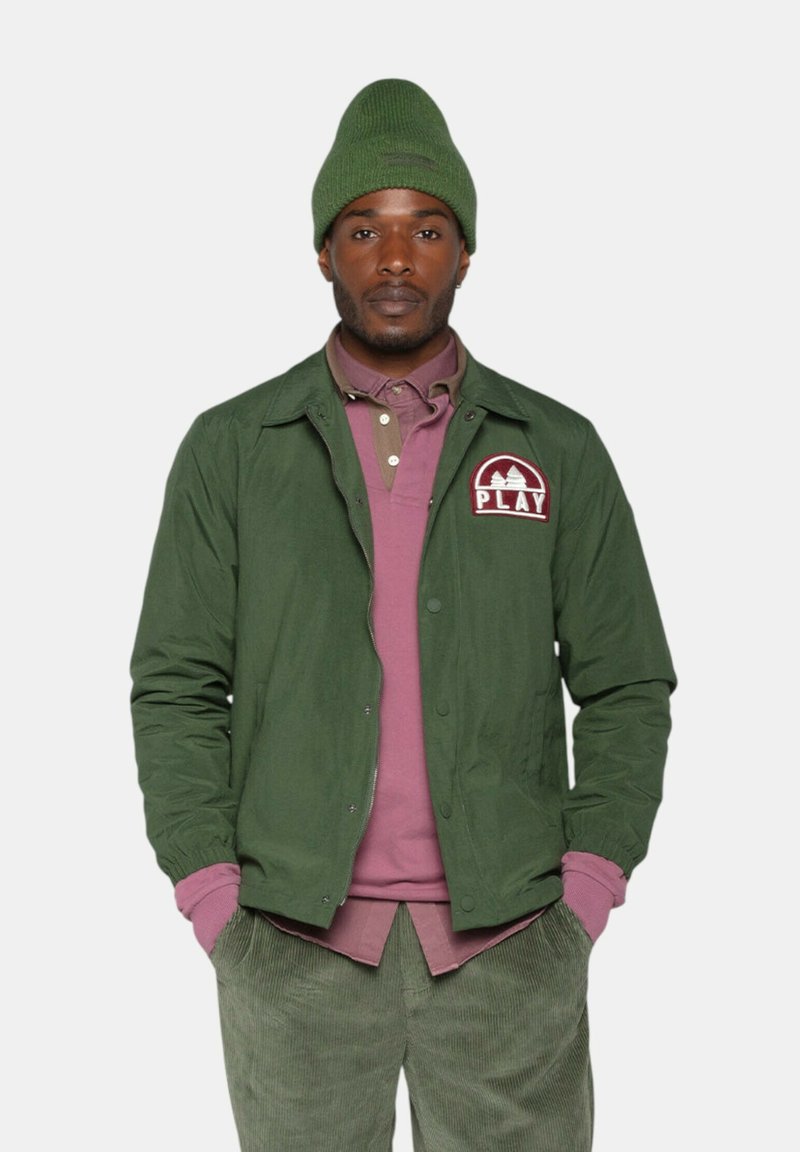 Veste verte avec logo, superposée sur une chemise violet clair. Texture lisse ; pantalon en velours côtelé de couleur verte atténuée. Bonnet en tricot vert.