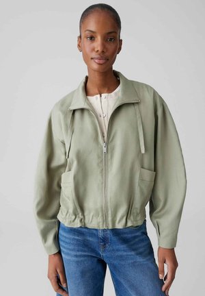 Donna con giacca verde chiaro con zip sopra una camicia bianca e jeans blu, in piedi davanti a uno sfondo grigio chiaro uniforme.