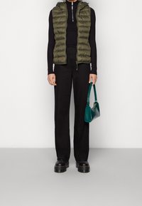 Chaleco acolchado verde oliva con capucha sobre un suéter negro de canalé, combinado con pantalones de pierna ancha negros y un bolso teal con un acabado texturizado.