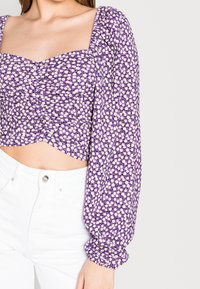 Fialový květinový top s nabíranými rukávy, zdobený malými bílými a žlutými květinovými vzory a čtvercovým výstřihem, doplněný bílými džíny.