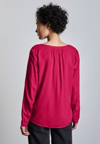 Blusa de manga larga en rosa brillante, hecha de un tejido suave. Presenta un escote redondo, detalle fruncido en la parte posterior y puños elásticos.
