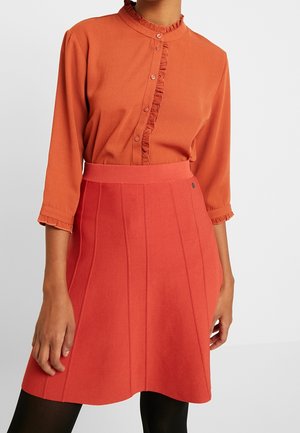 Femme portant un chemisier orange rouille à manches longues avec des volants et une jupe rouge corail en ligne A avec des coutures verticales.