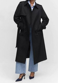 Czarny płaszcz trencz o fasonie dwurzędowym, z dużymi klapami, pagonami na ramionach i bocznymi kieszeniami, noszony na jasnoniebieskiej koszuli i dżinsach.